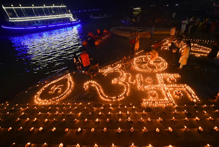 Thousands of lit diyas arranged into Om motifs and Hindi lettering on the ghat steps at night, a lit boat on the dark river behind