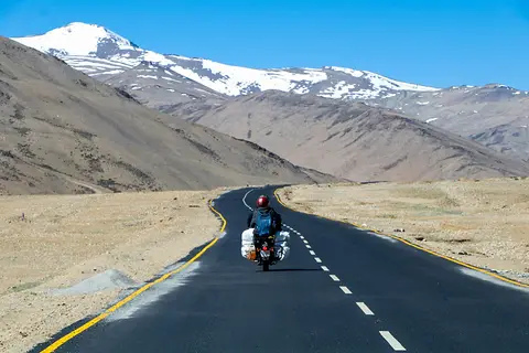 A motorcyclist with luggage riding a winding mountain road toward snow-capped peaks