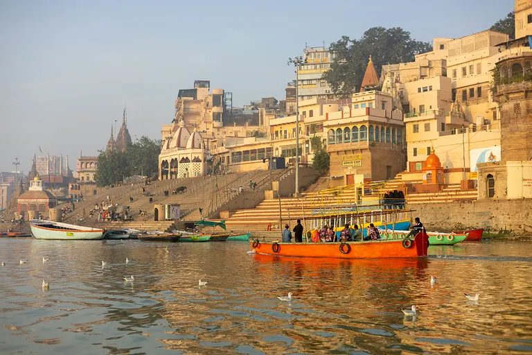 An orange sightseeing boat crossing the Ganga below the ghats in golden morning light, gulls on the water